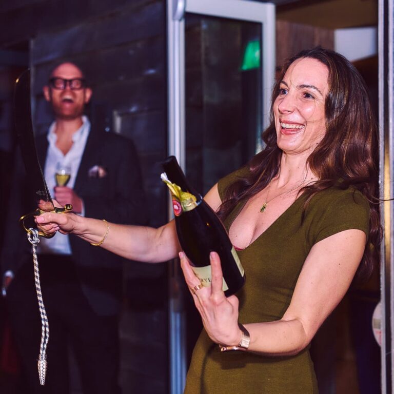 Woman in an olive dress holding a sparkling wine bottle and sabre