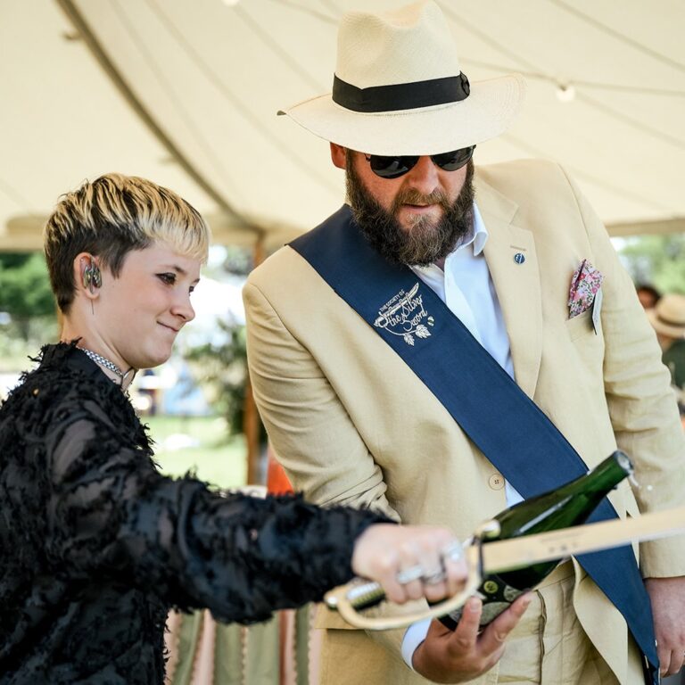 Man in a cream suit and sash guiding a guest through sabrage in a marquee