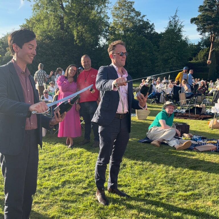Man performing sabrage in front of seated guests on a lawn