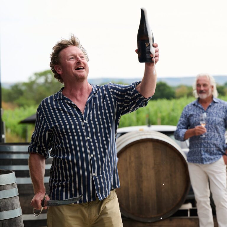 Man raising a sparkling wine bottle beside a barrel in a vineyard