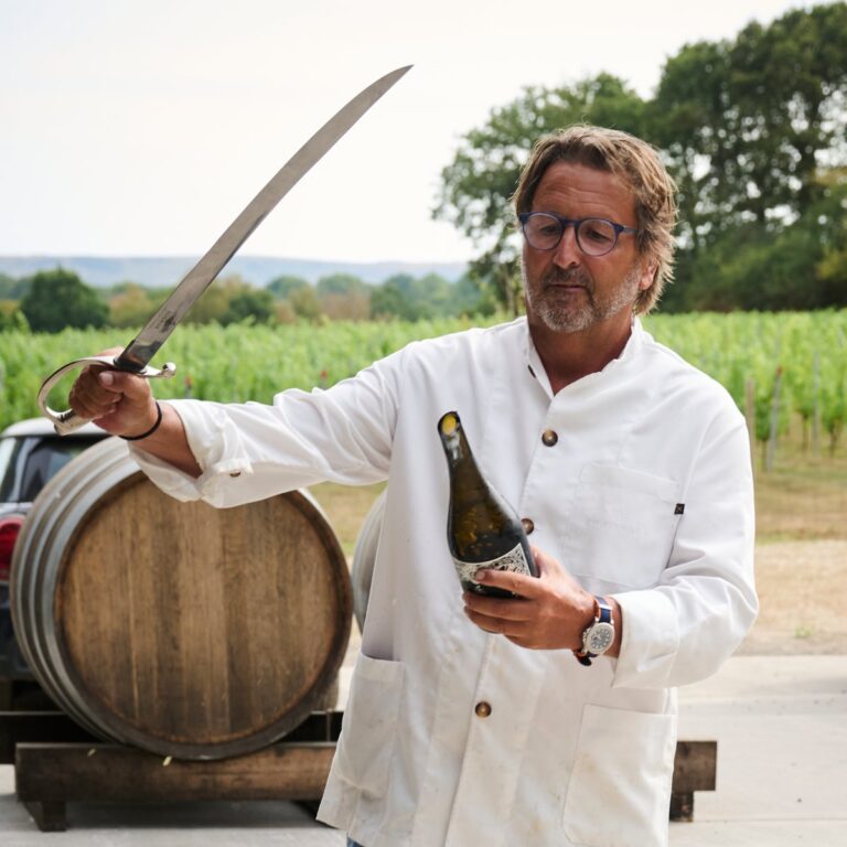 Man in white beside a barrel holding a sparkling wine bottle and sabre