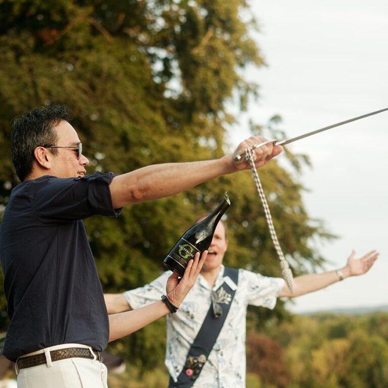 Man performing sabrage on a bottle held by another guest