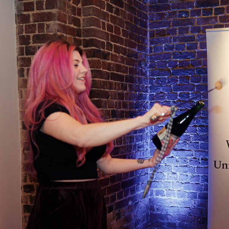 Woman with pink hair performing sabrage by a brick wall