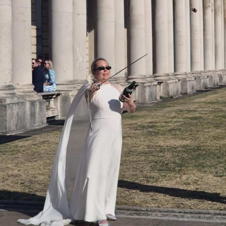 Woman in a long white dress performing sabrage in front of stone columns